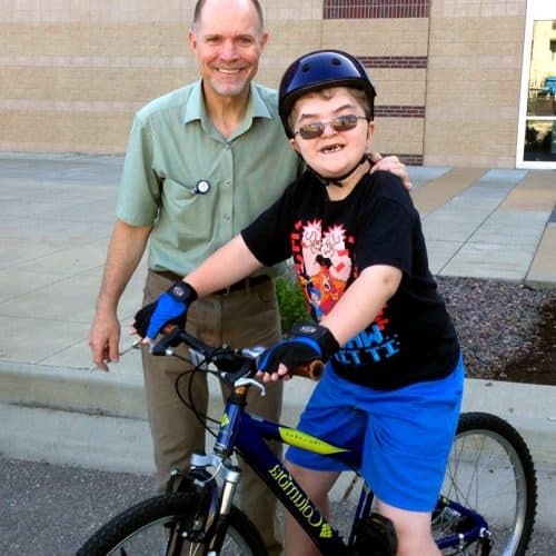 A smiling man in a green shirt stands beside a boy in a helmet and sunglasses sitting on a blue bike.