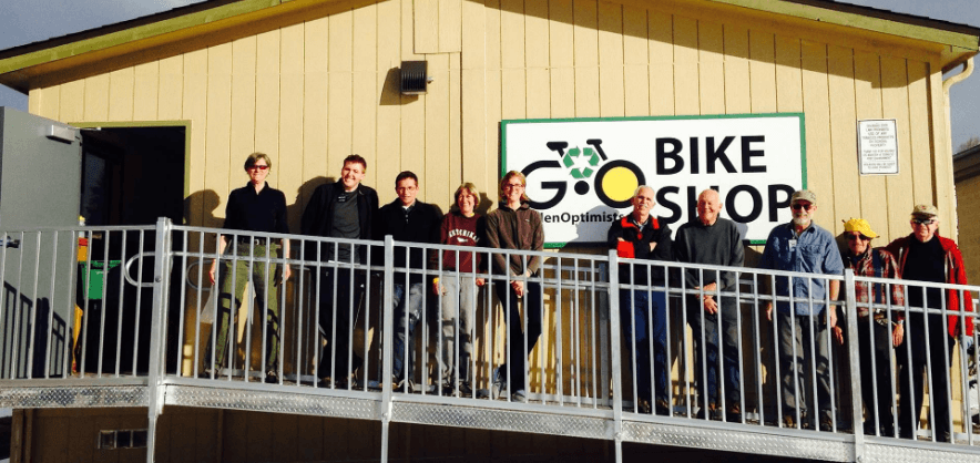 Ten Golden Optimists volunteers standing on the ramp in front of the tan bike shop, under the G·O BIKE SHOP sign.